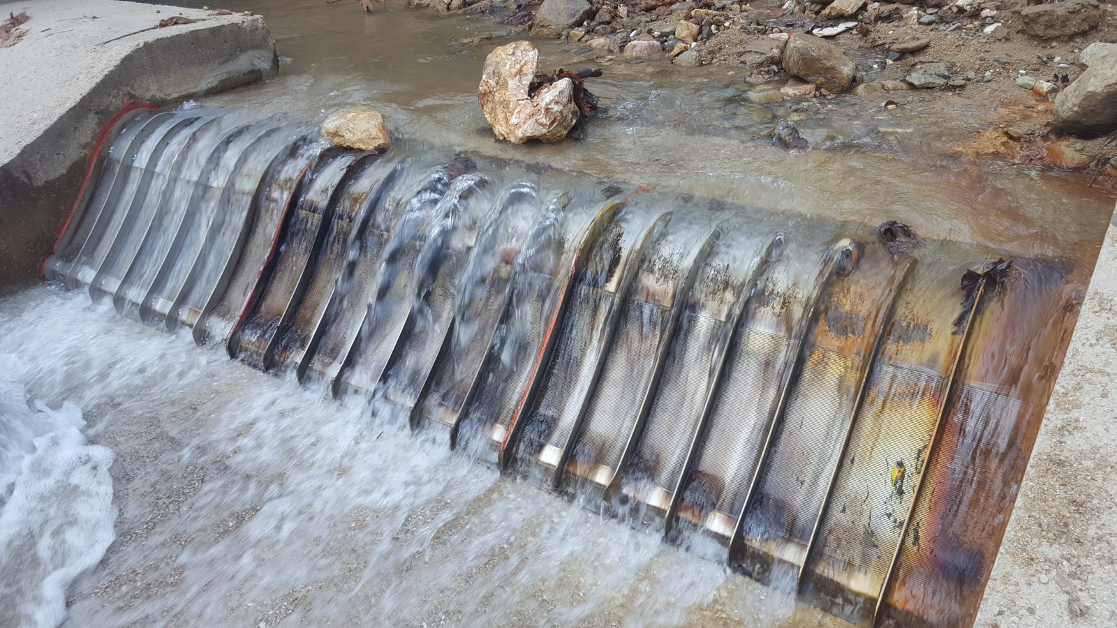 The same intake at the end of August 2018, after the flood: the pond behind the weir filled to the weir crest with rock and sand, boulders resting on the screen, water still flowing through the wedge wire